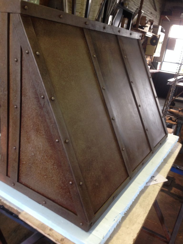 Kitched hood made from sheets of steel held together with rivets to a flat bar frame.  Patina made with vinegar, and hand buffed.