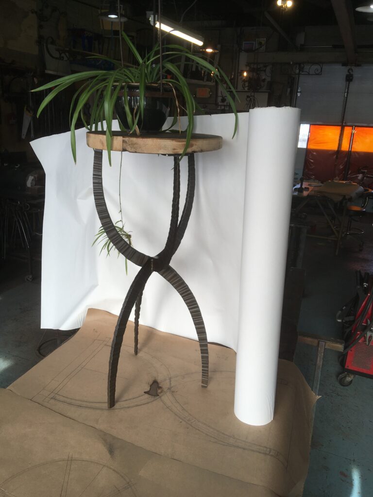 Forged end table with a plant on top, photographed in a metal shop