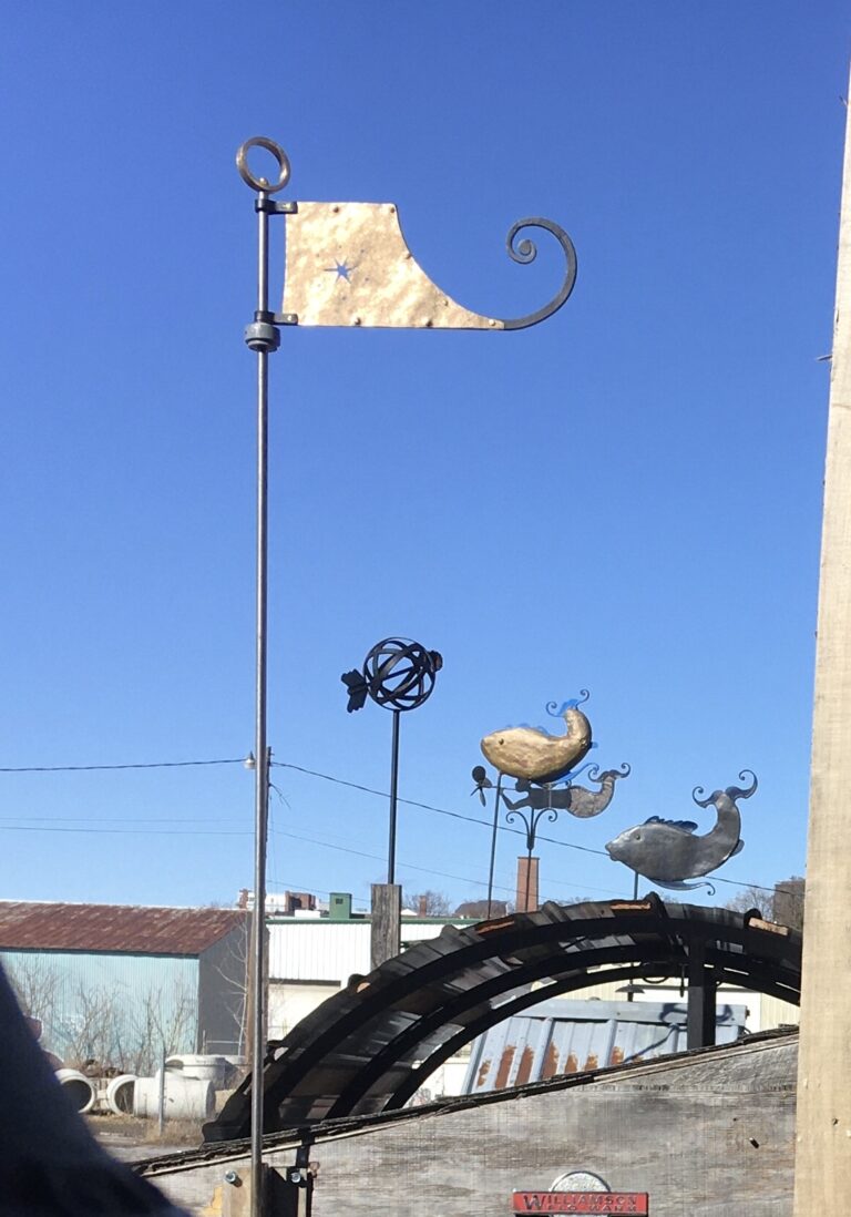 Flag finial on a tall pole with weathervanes on a rooftop behind it