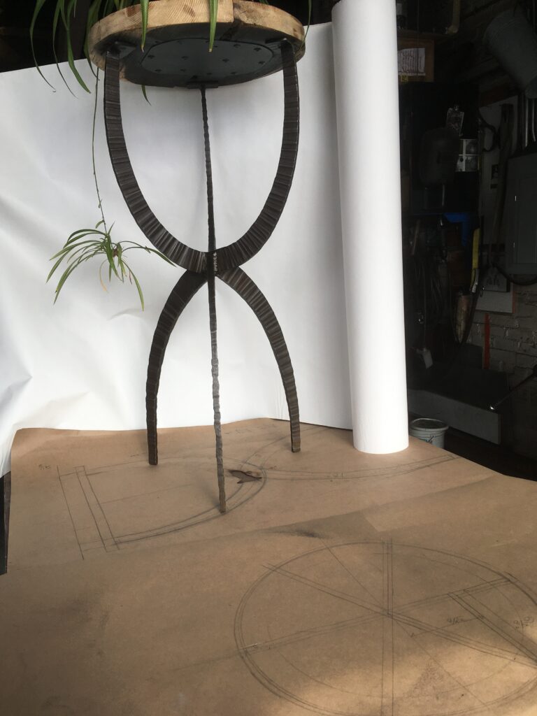 Forged metal end table with an hourglass base and a round top holding a plant