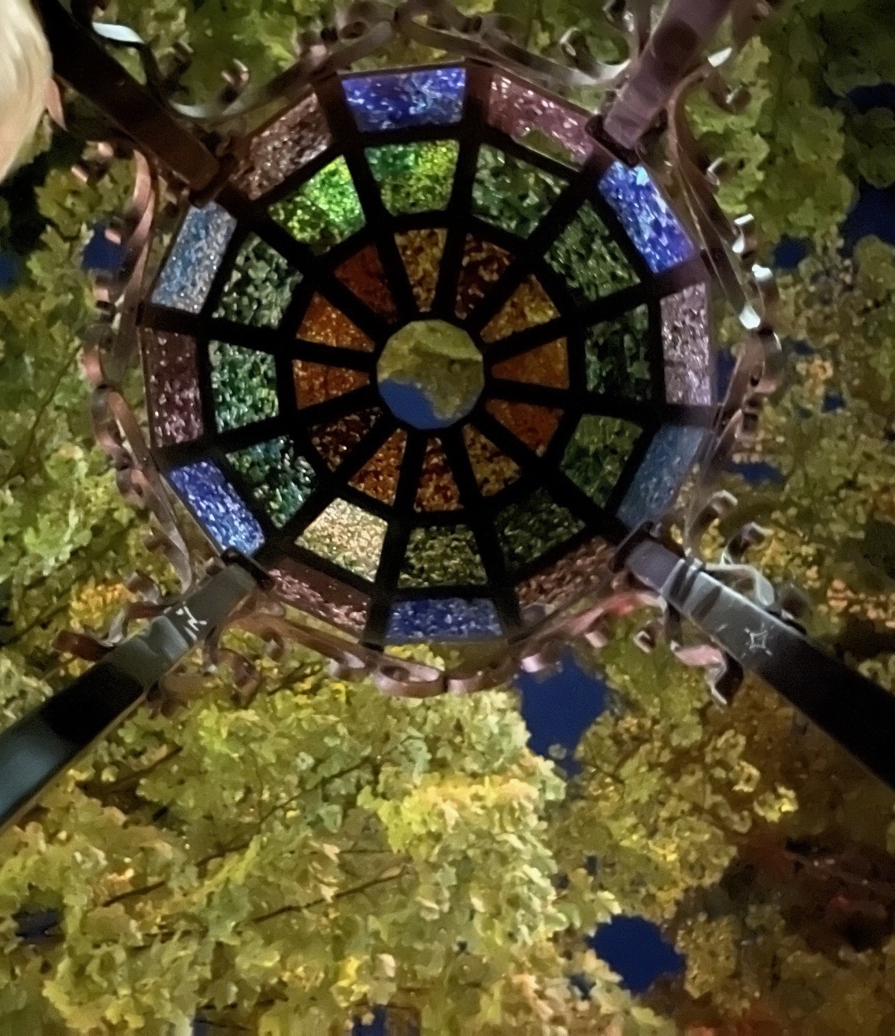 Looking up at a round stained glass dome set in a metal frame, with trees beyond